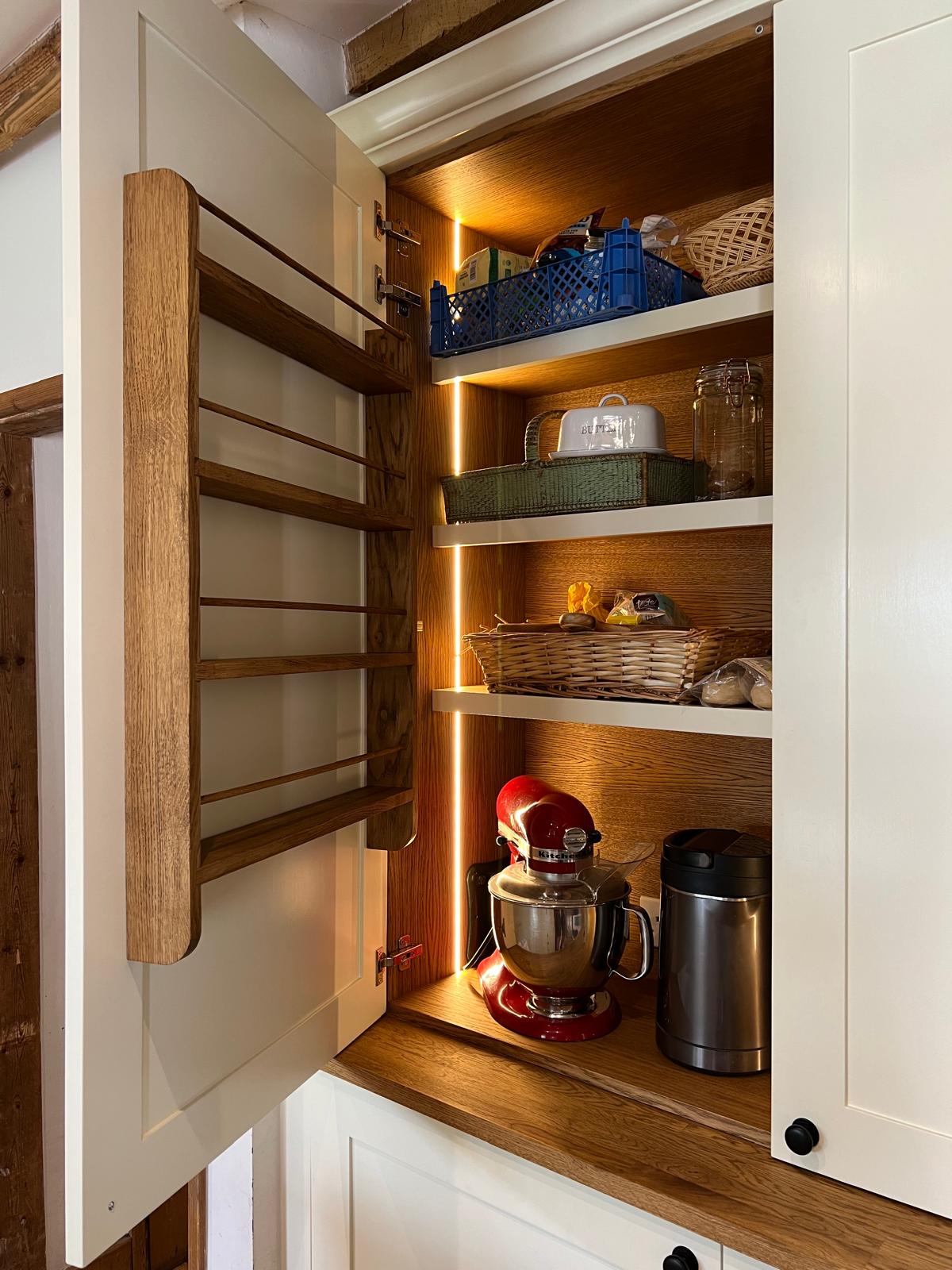 Fitted larder pantry open to show oak-lined shelving, spice racks and lighting